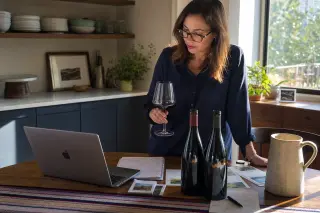 Woman in a sunlit home kitchen, thoughtfully comparing notes and wine bottles with a glass in hand, preparing to buy wine online.