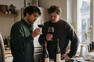 Two friends in a cozy kitchen compare two glasses of red wine by natural daylight, bottles with different labels subtly visible, evoking an informal, thoughtful wine discussion.