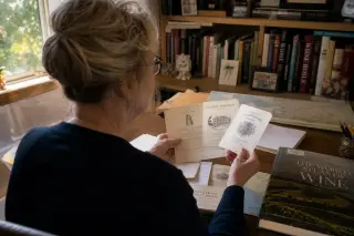 Woman in a casual home office gently examining Bordeaux wine labels at her desk, sunlight filtering in, with notes and books nearby