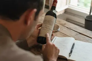 Person at home examining a French wine bottle label, sunlight highlighting handwritten notes and offering a sense of discovery.